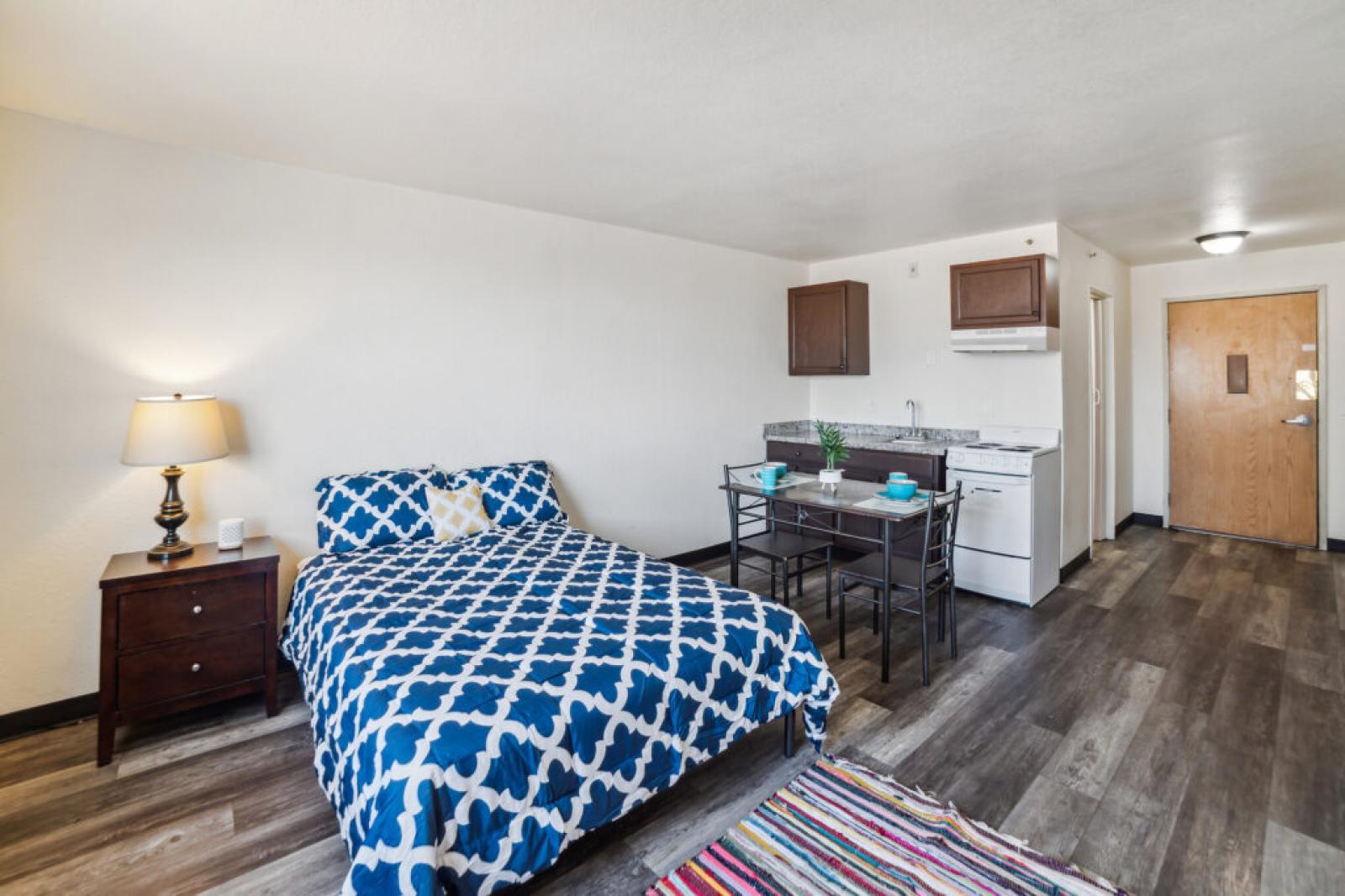 Compact studio apartment with a bed featuring blue and white patterned bedding, a small dining table, and a kitchenette.