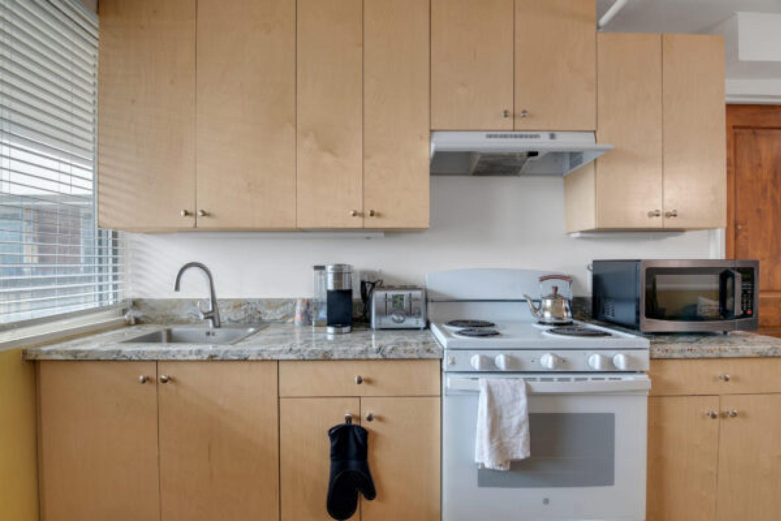 Modern kitchen with light wood cabinets, granite counters, white stove, microwave, and sink by a window.
