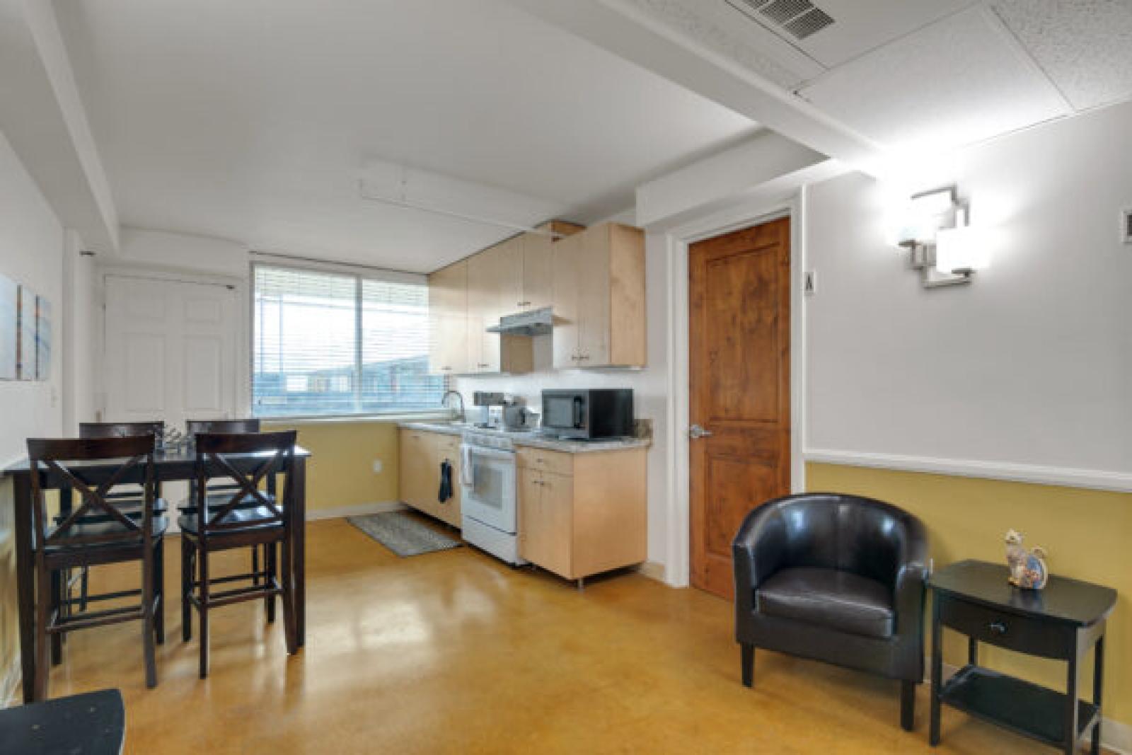Open-plan kitchen and dining with light wood cabinets, dark dining set, yellow floor, and a brown leather armchair.
