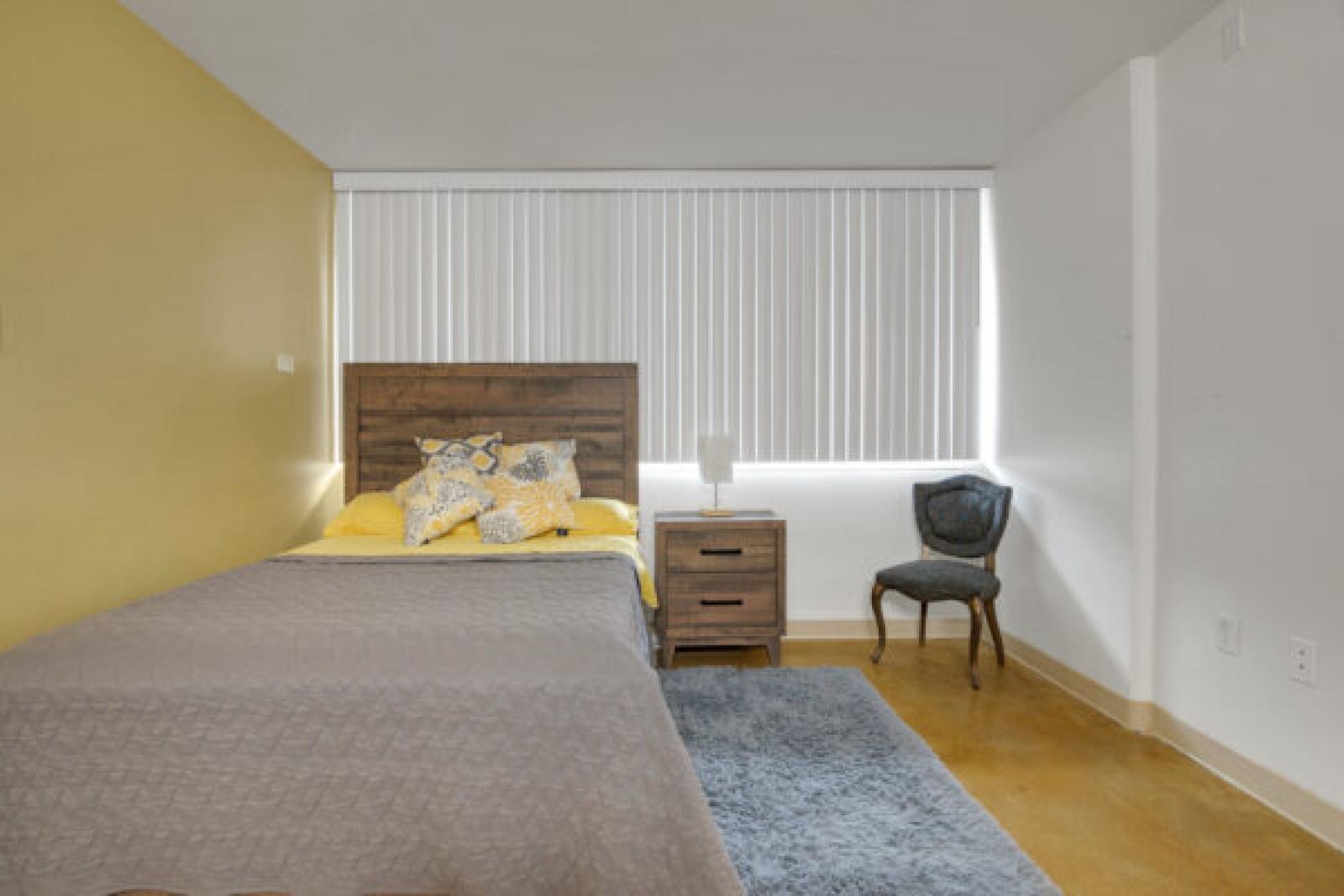 Bedroom with a single bed, wooden headboard, yellow accent wall, and white walls. A nightstand and dark chair sit by the window with blinds.