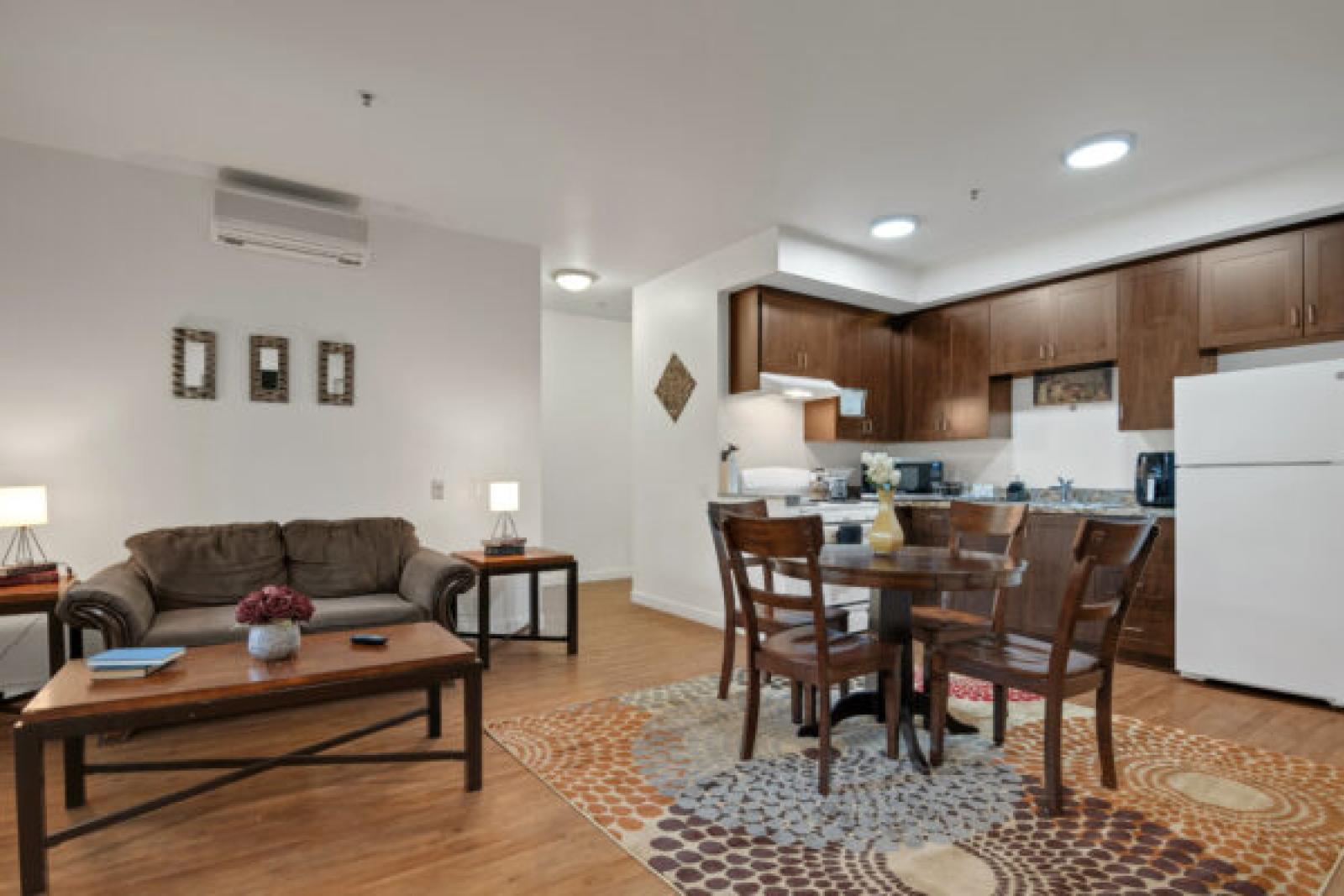 Modern apartment: living room with brown sofa, dining area with round table, and dark wood kitchen cabinets.