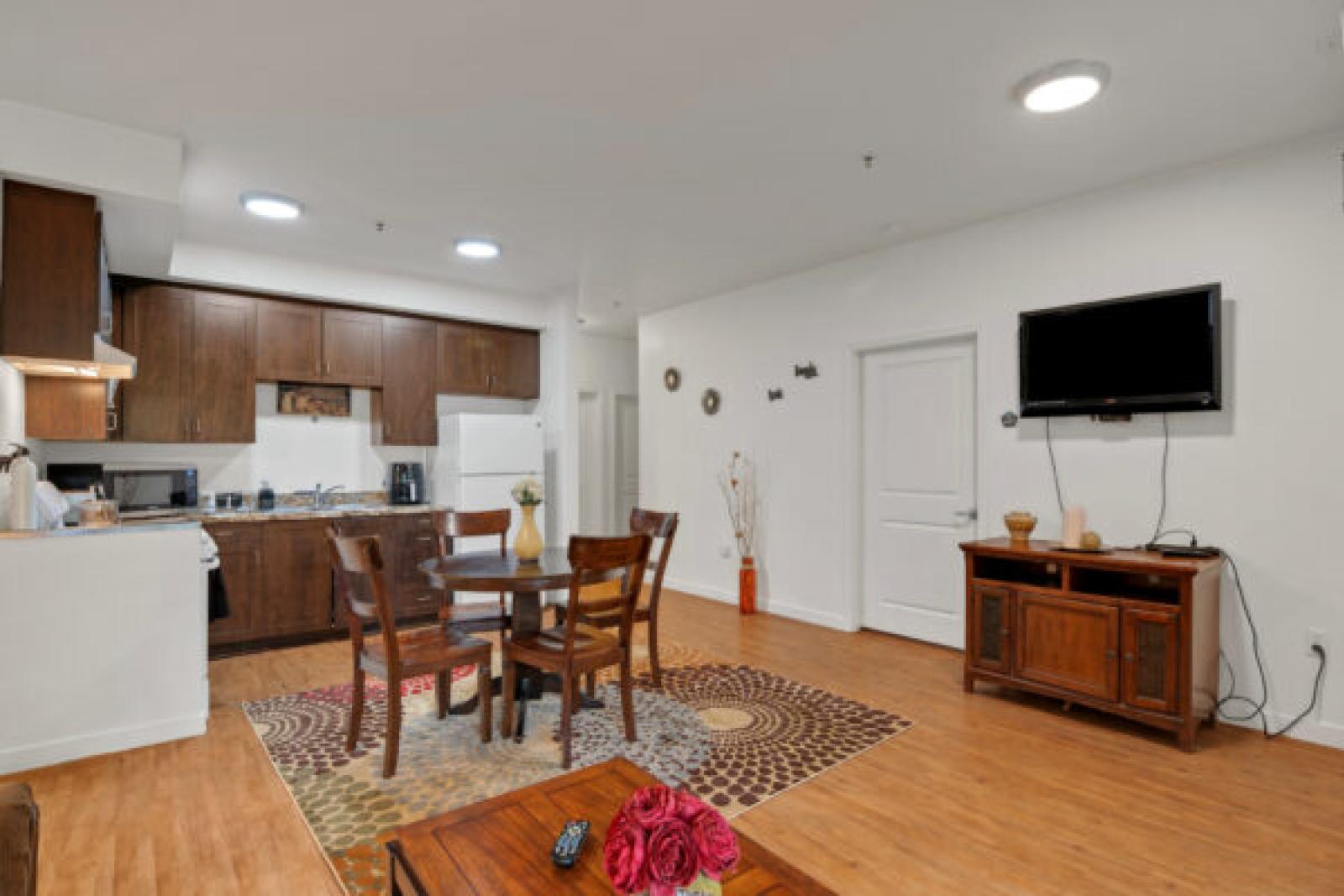 Open-plan apartment with a dark wood kitchen, round dining table on a patterned rug, and a living area with a TV.