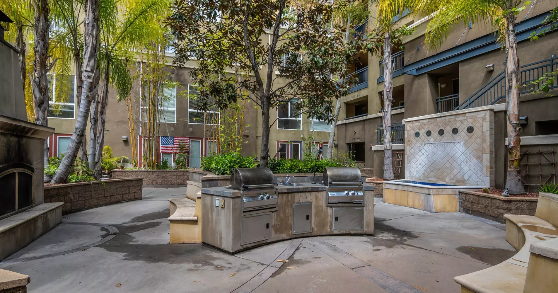 Paved outdoor courtyard with built-in BBQ grills, palm trees, and a wall fountain next to apartment buildings.