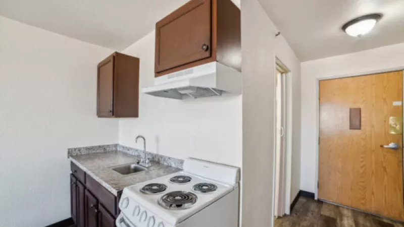 Compact kitchen with dark brown cabinets, white electric stove, and speckled counter, next to an entryway with a light brown door.