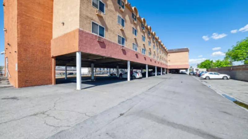 Apartment or hotel building with tan and brick-red facade, offering shaded ground-floor parking for vehicles under a clear blue sky.