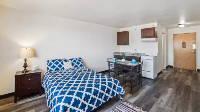 Studio apartment with a blue and white patterned bed, nightstand, dark wood-look floor, and compact kitchen area.