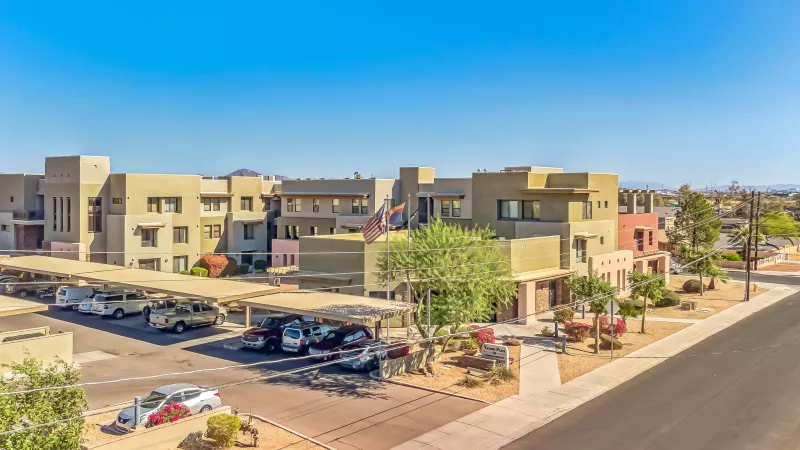 Aerial view of a modern tan apartment complex with colored accents, covered parking, and a clear blue sky.