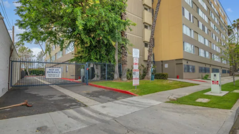 Tan multi-story apartment building behind a closed metal gate, with green trees, a sidewalk, and street.