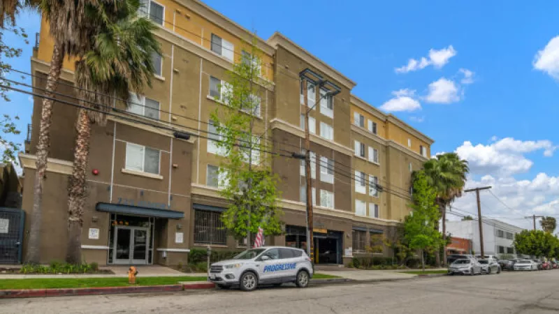 Modern multi-story apartment building with tan and brown facade, palm trees, and a street with vehicles.
