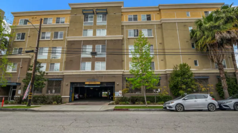 Multistory apartment building with cream and olive green facade, a parking garage entrance, street parking, and trees under a blue sky.
