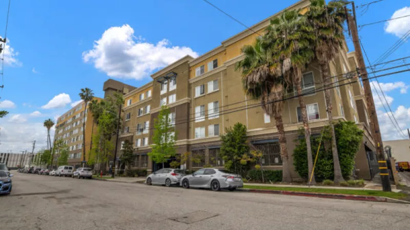 Large, multi-story building with cars parked on the street under a blue sky, flanked by palm trees.