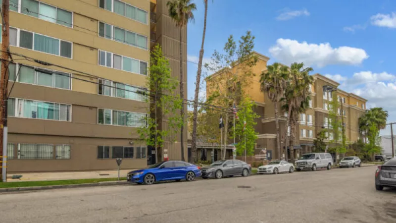 Street view with parked cars along a sidewalk next to multi-story buildings and palm trees under a cloudy sky.