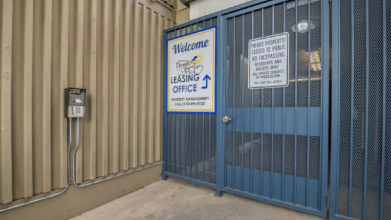 Blue metal gate with a "Welcome Leasing Office" sign and a "Private Property" sign, next to a tan corrugated wall.