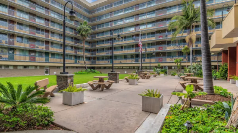 Courtyard with a green lawn, concrete paths, picnic tables, benches, palm trees, and a multi-story building in the background.