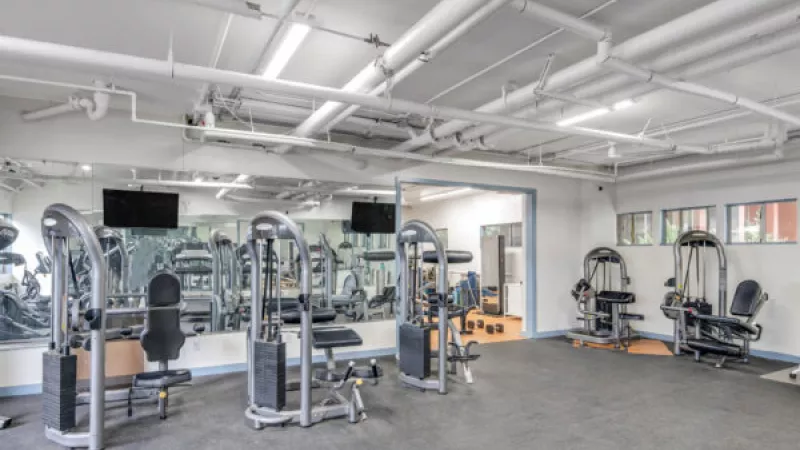Gym with various weight machines, a mirrored wall, and exposed white pipes on the ceiling.