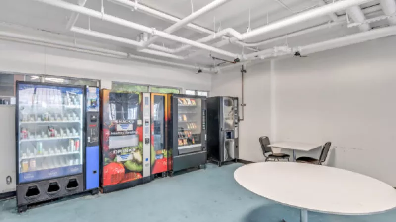 Four vending machines line a white wall in a breakroom with a blue carpet and two white tables.