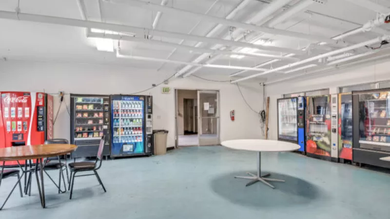 Vending room with several snack and drink machines, two round tables, and a doorway. Fluorescent lights illuminate the blue floor.