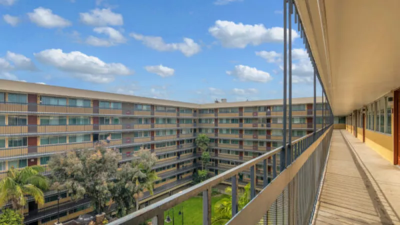 Outdoor building walkway overlooking a multi-story courtyard with green grass, trees, yellow walls, and a cloudy blue sky. Photograph.