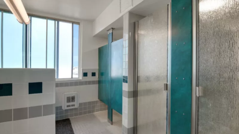 Public shower room with teal and white stalls, frosted glass doors, white tiled walls, and a large window.