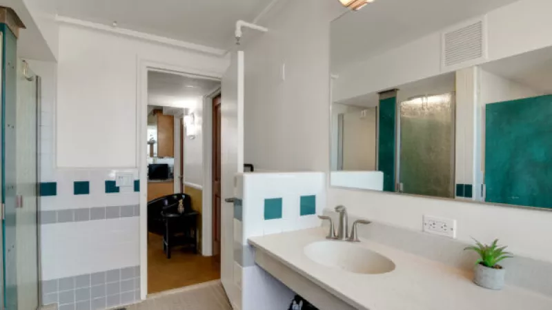 Bright bathroom with a white vanity, sink, large mirror, and teal accent tiles. A glass shower is visible on the left.