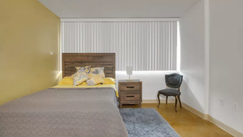 Bedroom with a single bed, wooden headboard, yellow accent wall, and white walls. A nightstand and dark chair sit by the window with blinds.
