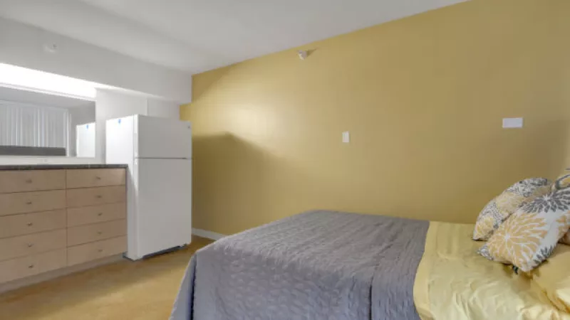 Studio apartment with a bed with grey comforter and yellow pillows, next to a kitchenette with white fridge and wood cabinets.