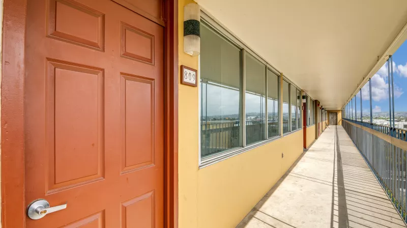 Long outdoor apartment hallway with an orange door, yellow walls, and windows reflecting the sky. Metal railing on right.