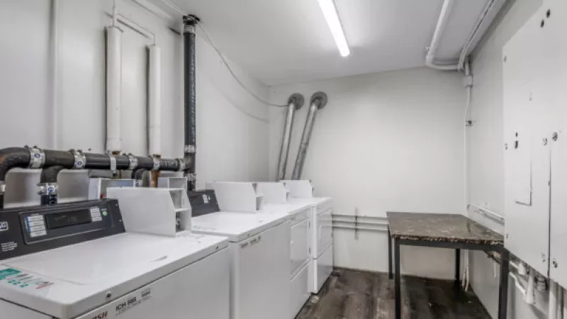White top-loading washing machines line a small laundry room with exposed pipes, dark wood-look floor, and a folding table.