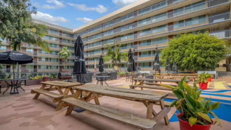 Outdoor patio with wooden picnic tables, black umbrellas, and potted plants in front of a large building.