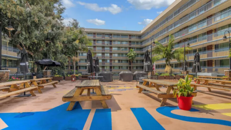 Outdoor courtyard with wooden picnic tables, black umbrellas, and colorful blue and yellow ground patterns, surrounded by a building.