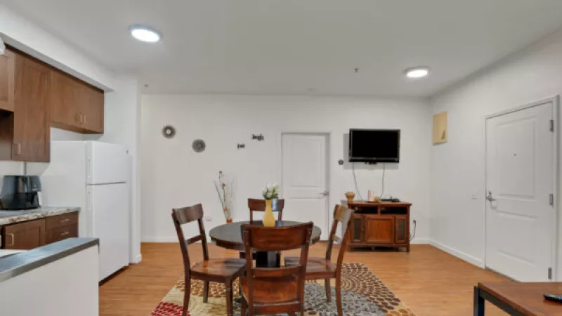 Open-concept living space with a kitchen, dining table with four chairs on a patterned rug, and a wall-mounted TV.