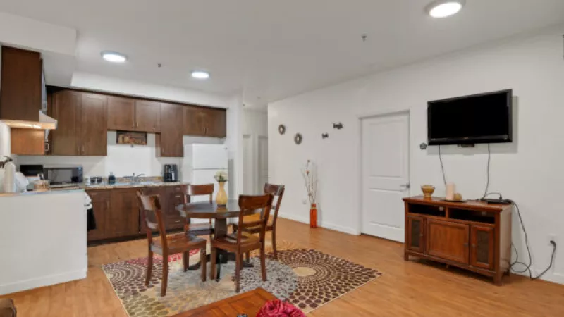 Open-plan apartment with a dark wood kitchen, round dining table on a patterned rug, and a living area with a TV.