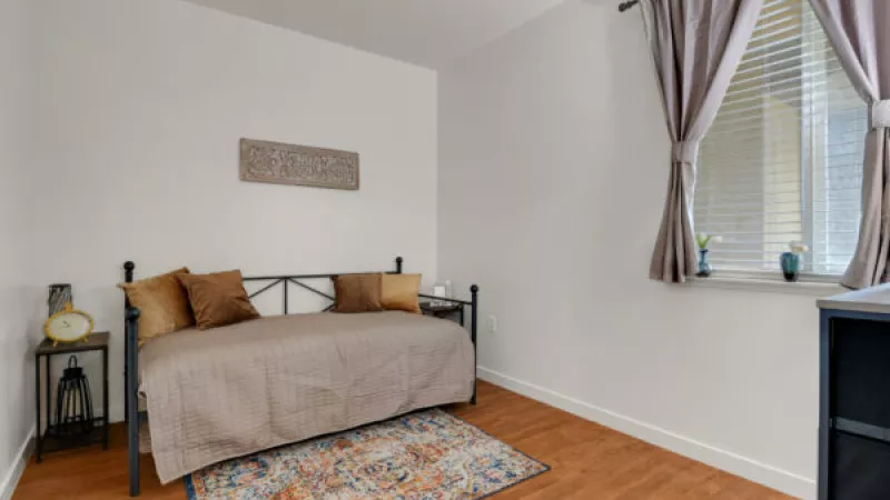 Small bedroom with a daybed, brown cushions, wood floor, and a patterned rug. A window with beige curtains.
