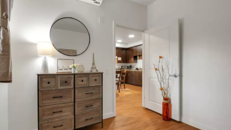 Interior room with a wooden dresser, round mirror, and lamp. An open door reveals a kitchen. An orange vase with branches stands nearby.