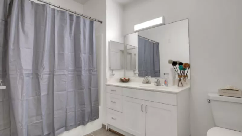 Modern bathroom with white vanity, two sinks, large mirror, grey shower curtain, and a toilet.