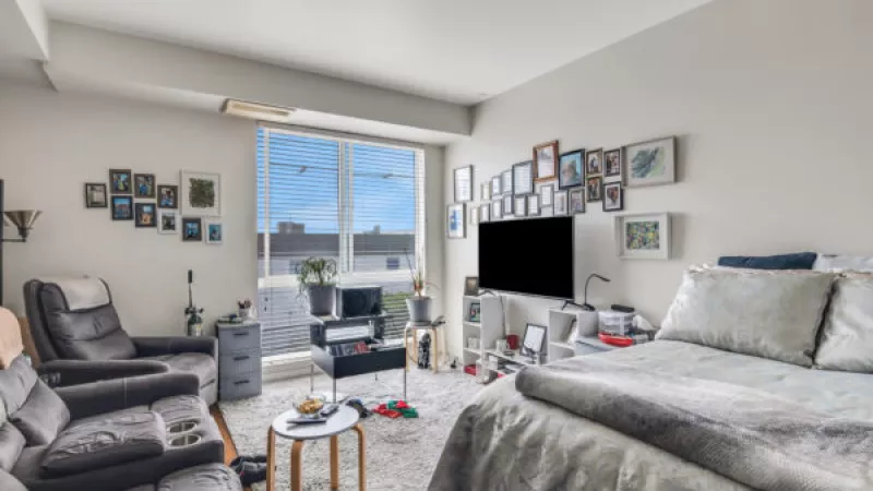 Bright studio apartment with a grey bed, sofa, and TV. Walls are covered in many framed photos, and a large window shows the sky.