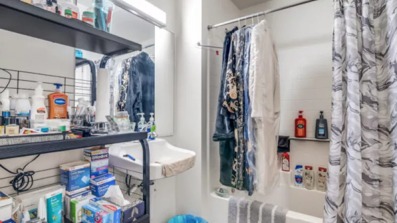 Small, cluttered bathroom with black shelves of toiletries, a small sink, and a shower with hanging robes and a patterned curtain.