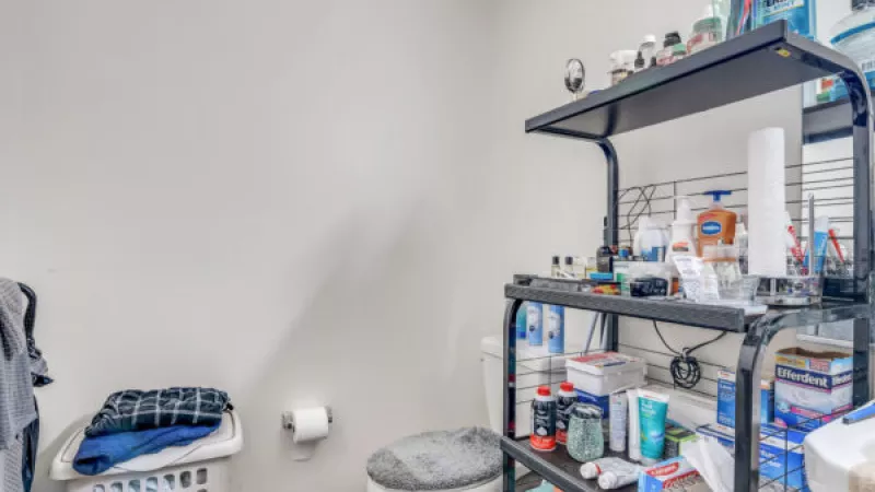 Small bathroom with a toilet, laundry hamper, and an overloaded black shelving unit filled with toiletries.