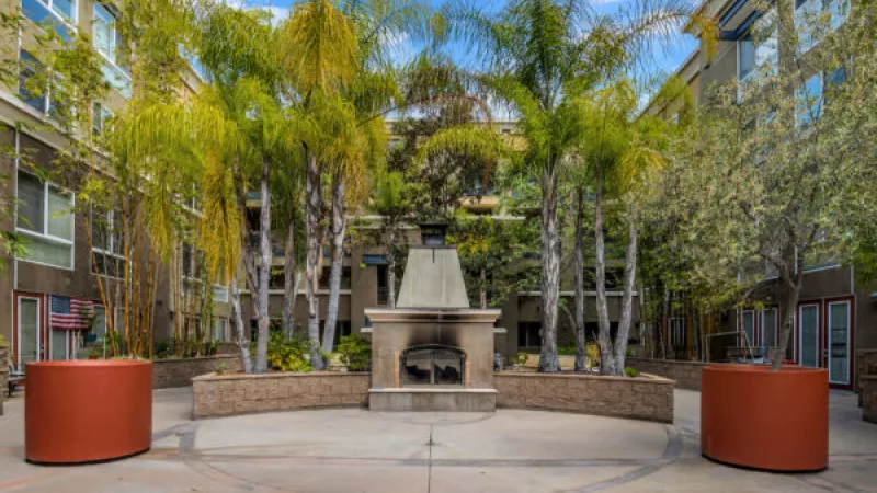 Outdoor courtyard featuring a central stone fireplace, surrounded by tall palm trees, green foliage, and residential buildings.