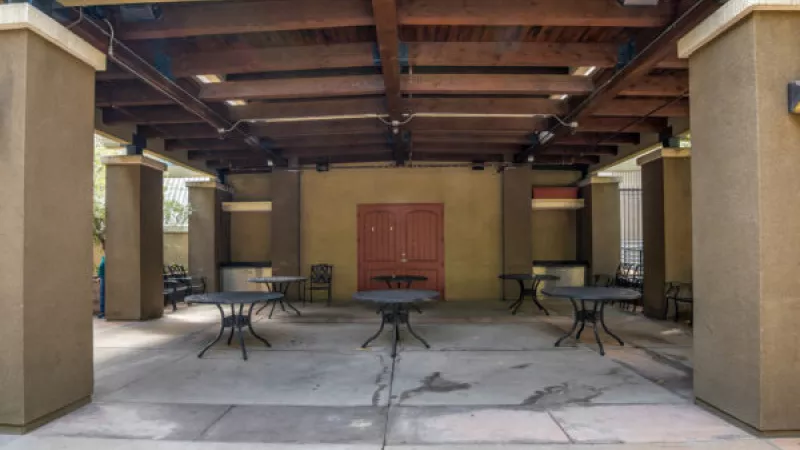 Outdoor covered patio with black tables, chairs, wooden beam ceiling, and a reddish-brown double door on a light brown wall.