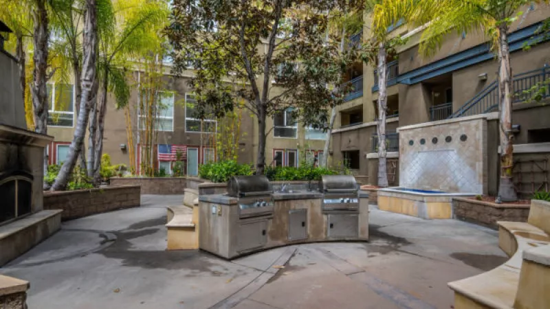 Modern apartment courtyard featuring a double barbecue station, tall palm trees, a waterfall, and outdoor seating.