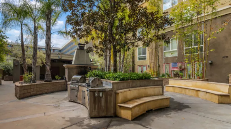 Outdoor courtyard with a large built-in BBQ grill, palm trees, leafy trees, and curved stone benches.