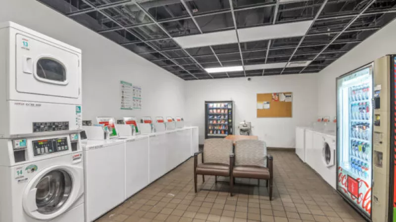Modern laundromat with many white washers and dryers, two vending machines, and a seating area with chairs and a table.