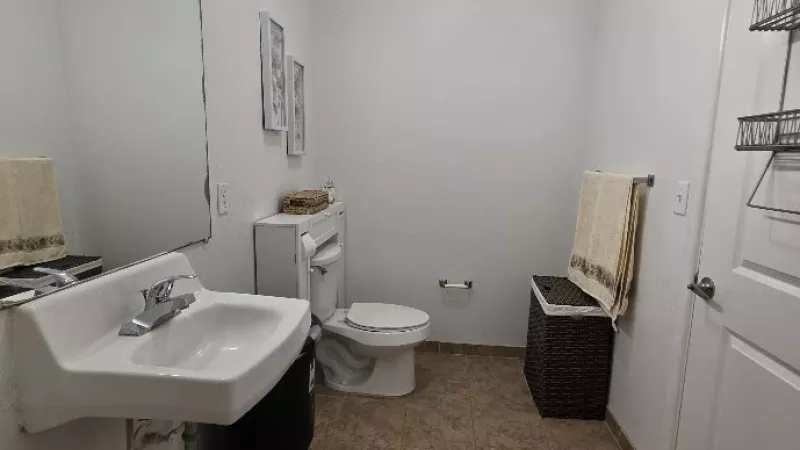 Bright bathroom with white wall-mounted sink, toilet, large mirror, and brown tiled floor. A dark bench sits near the door.