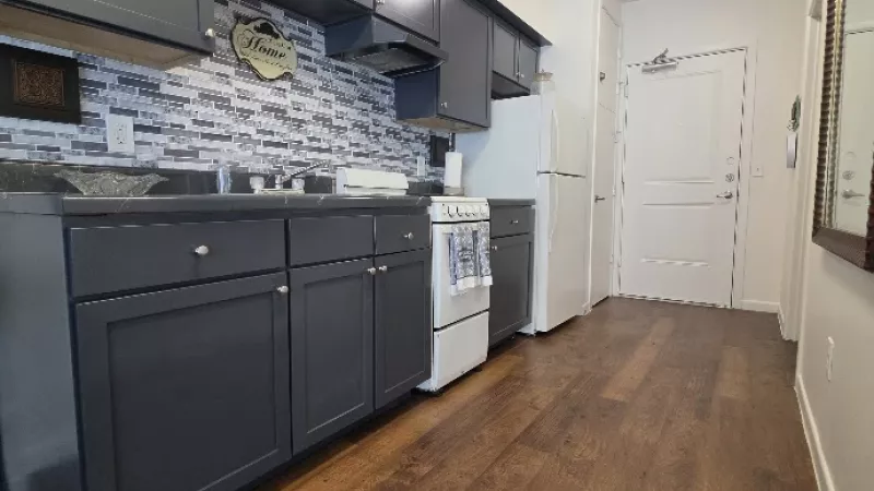 Modern kitchen with dark gray cabinets, white appliances, and gray-white tiled backsplash. Wood floor leads to a white door.