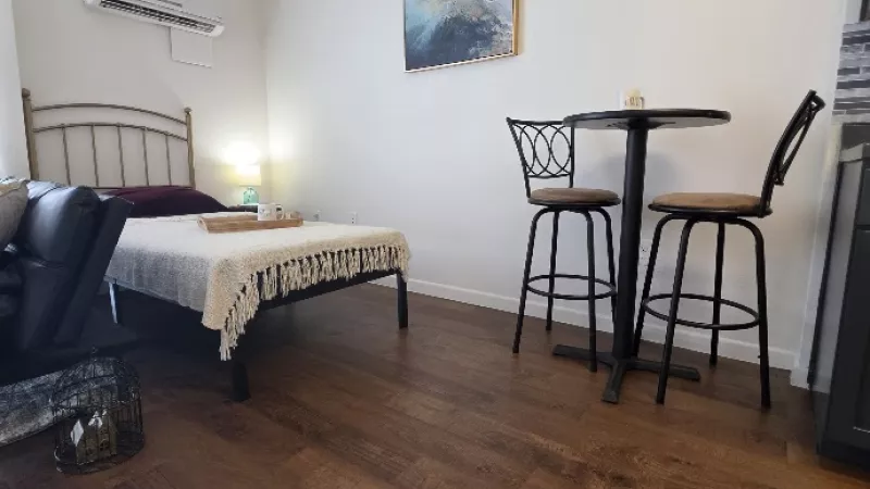 A well-lit studio room featuring a bed, small dining table, coffee table, abstract art, and rich wooden flooring.