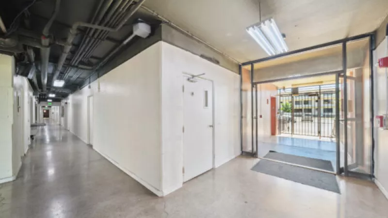 Long indoor hallway with white walls, exposed ceiling pipes, and an open glass door leading to an outdoor area.