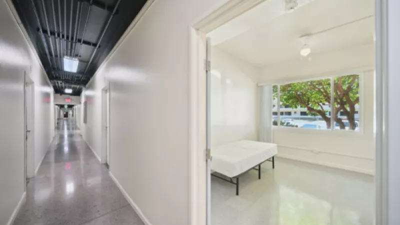 Photograph: Long white hallway with dark ceiling, exposed pipes, and an open door to a minimalist room with a bed and window.