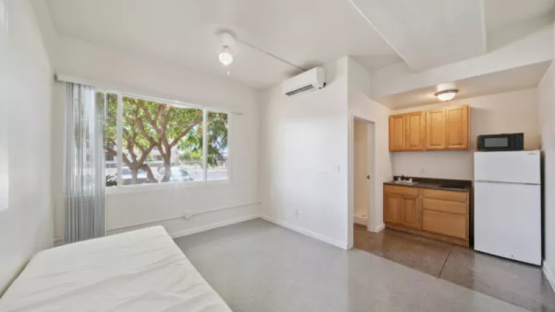 Small studio apartment with a mattress on the floor, large window, and a compact kitchenette featuring wood cabinets.