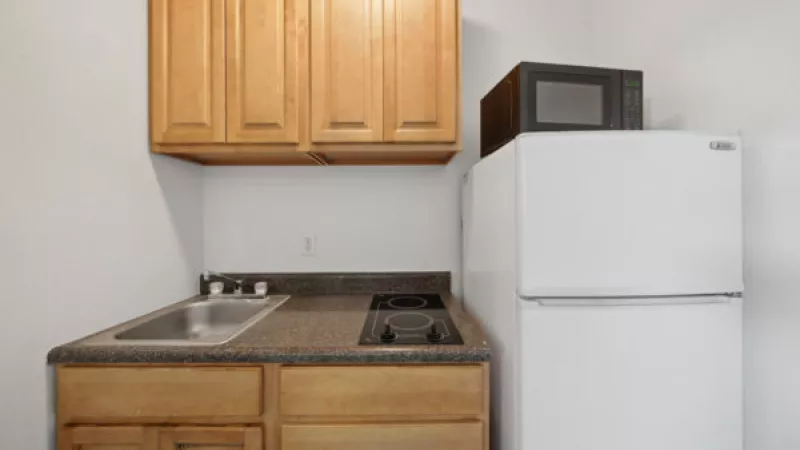Small kitchen with light wood cabinets, sink, two-burner cooktop, and white refrigerator with a microwave on top.